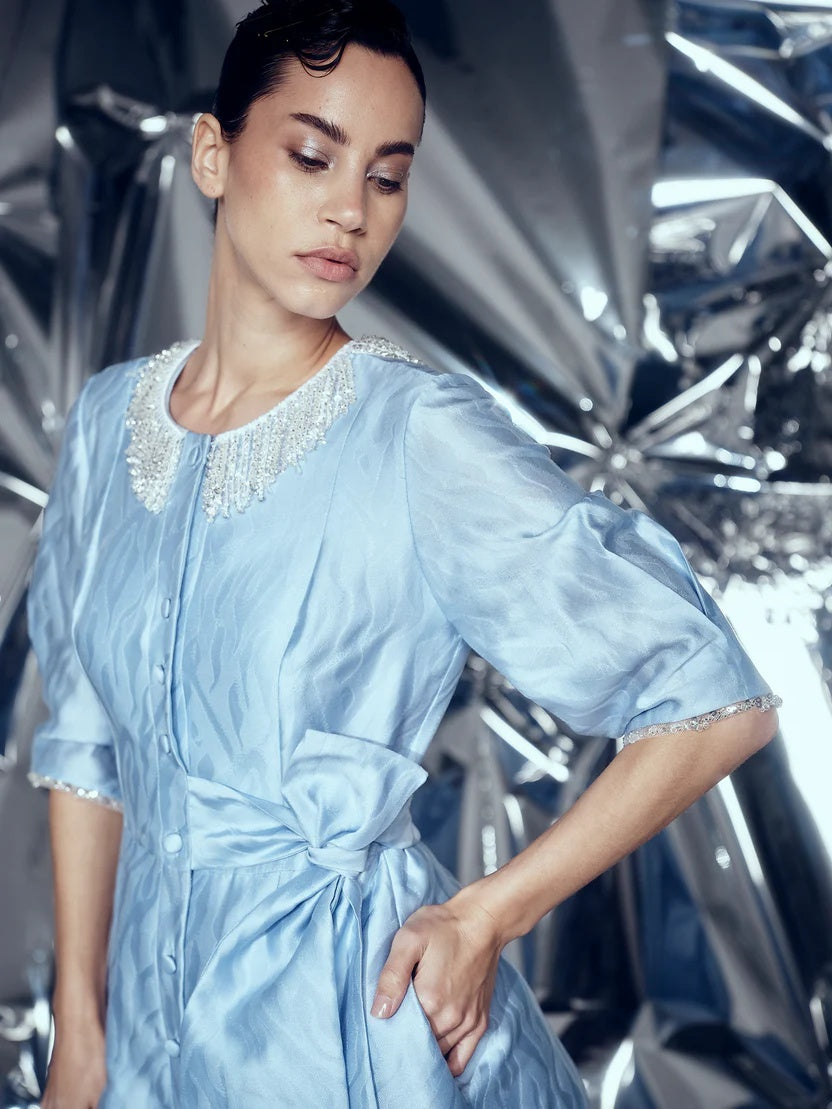 A woman wearing a light blue satin dress with a beaded crew neck and ruffled sleeves, posing against a reflective metallic silver backdrop.