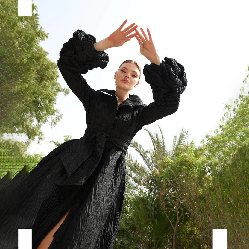 Woman in a black dress with ruffled sleeves standing outdoors with trees