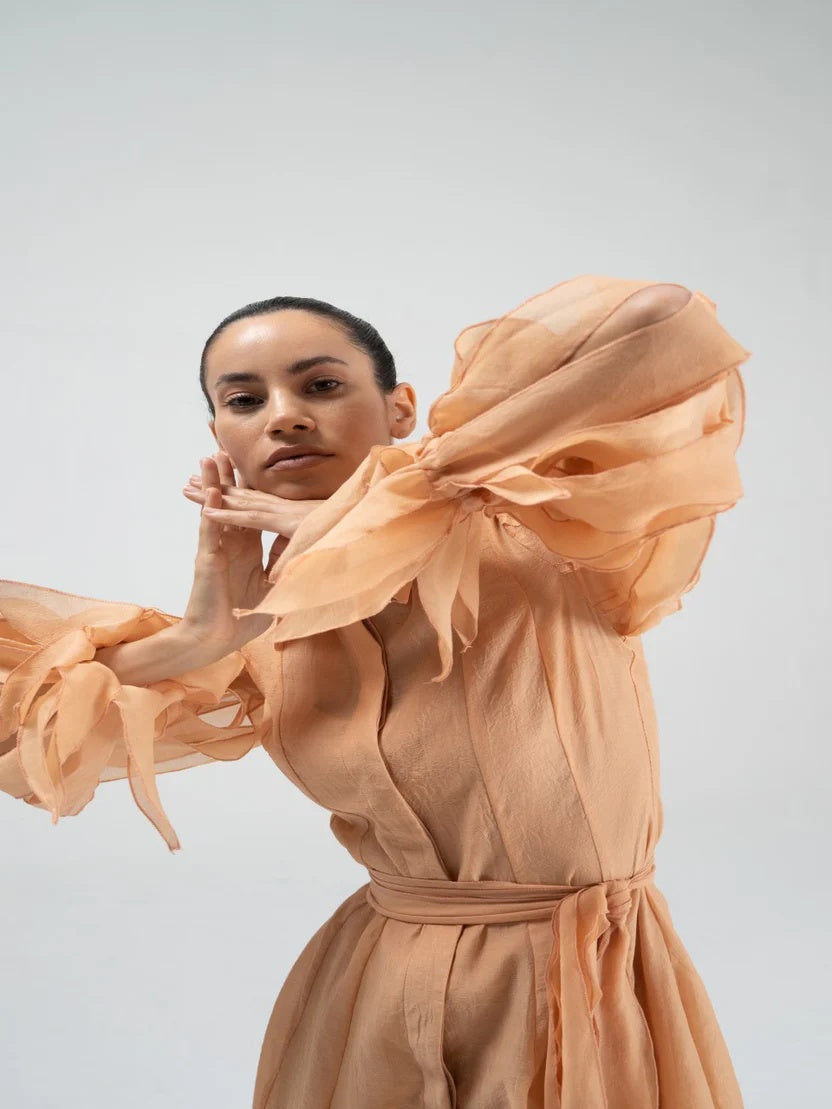 A model wearing a peach-colored dress with dramatic, voluminous sheer ruffle sleeves and a matching waist tie, posed against a light gray studio background.